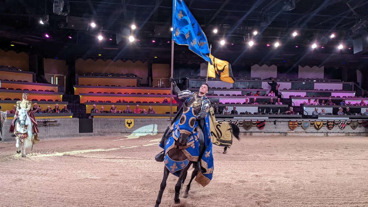 A knight carrying a blue and gold banner rides across the arena at Medieval Times, with the queen on a white horse and the crowd behind